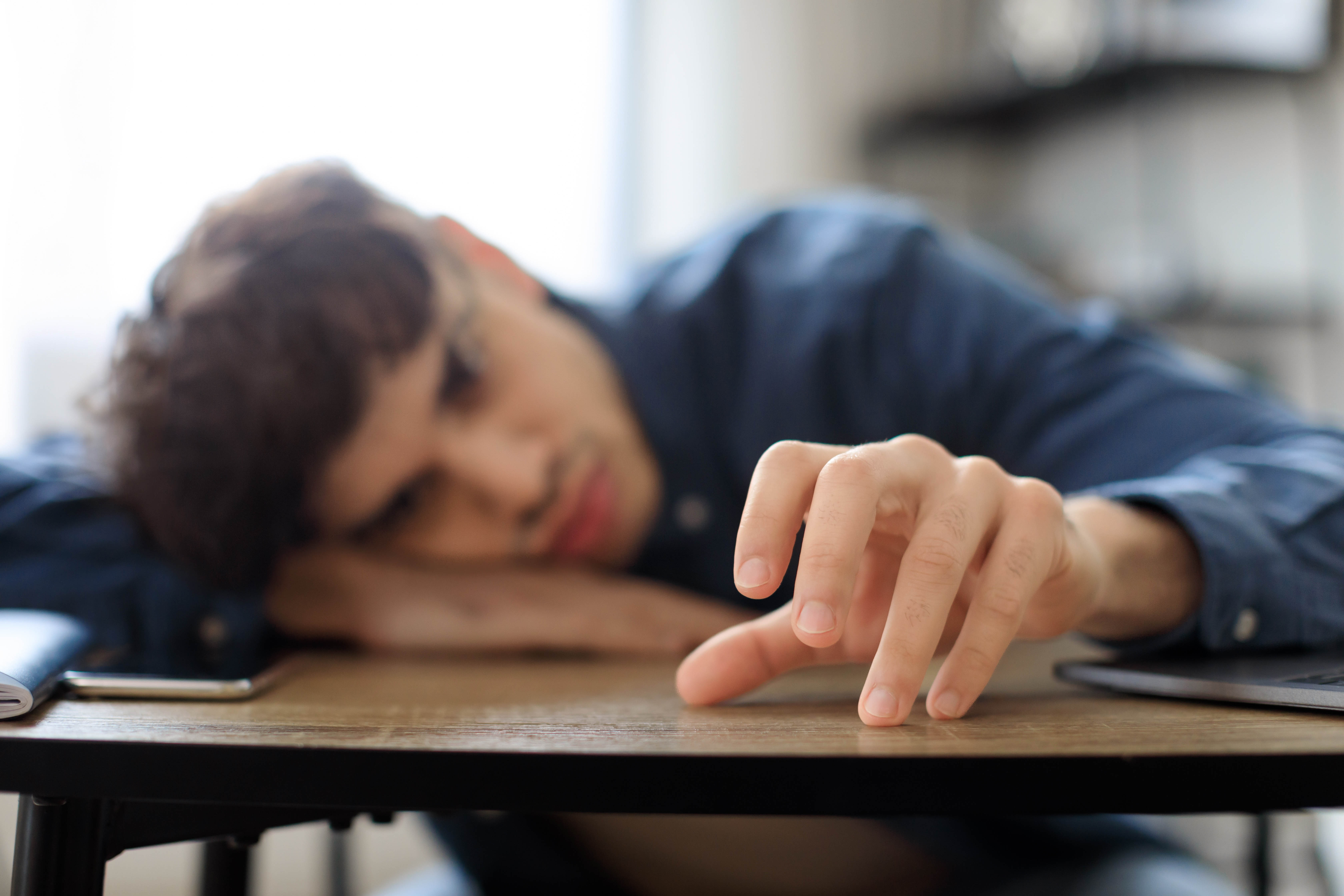 Man holding his hand in front of himself and looking at it intently, hand is drumming on a desk while the man has his head down and on its side on his other arm